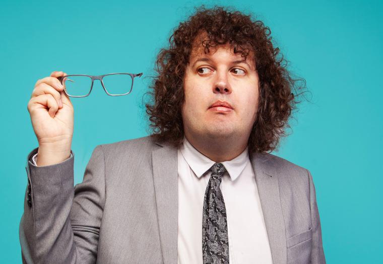 A man in a grey suit holds his glasses to one side - comedian Ray O'Leary