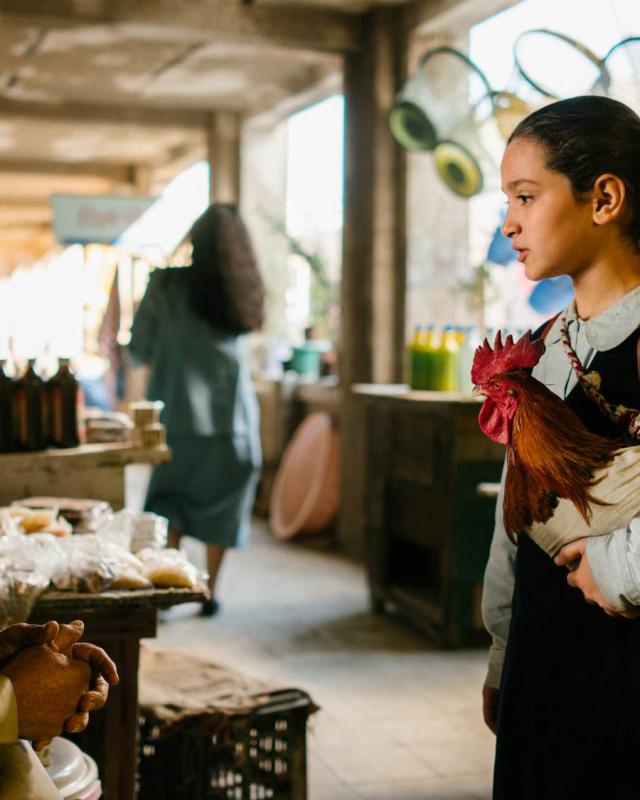 A young girl holds a rooster as she talks to a man in a market stall