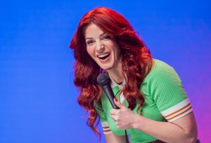 A woman with red hair, wearing a green dress, holds a microphone - Bron Lewis