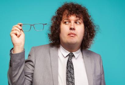 A man in a grey suit holds his glasses to one side - comedian Ray O'Leary